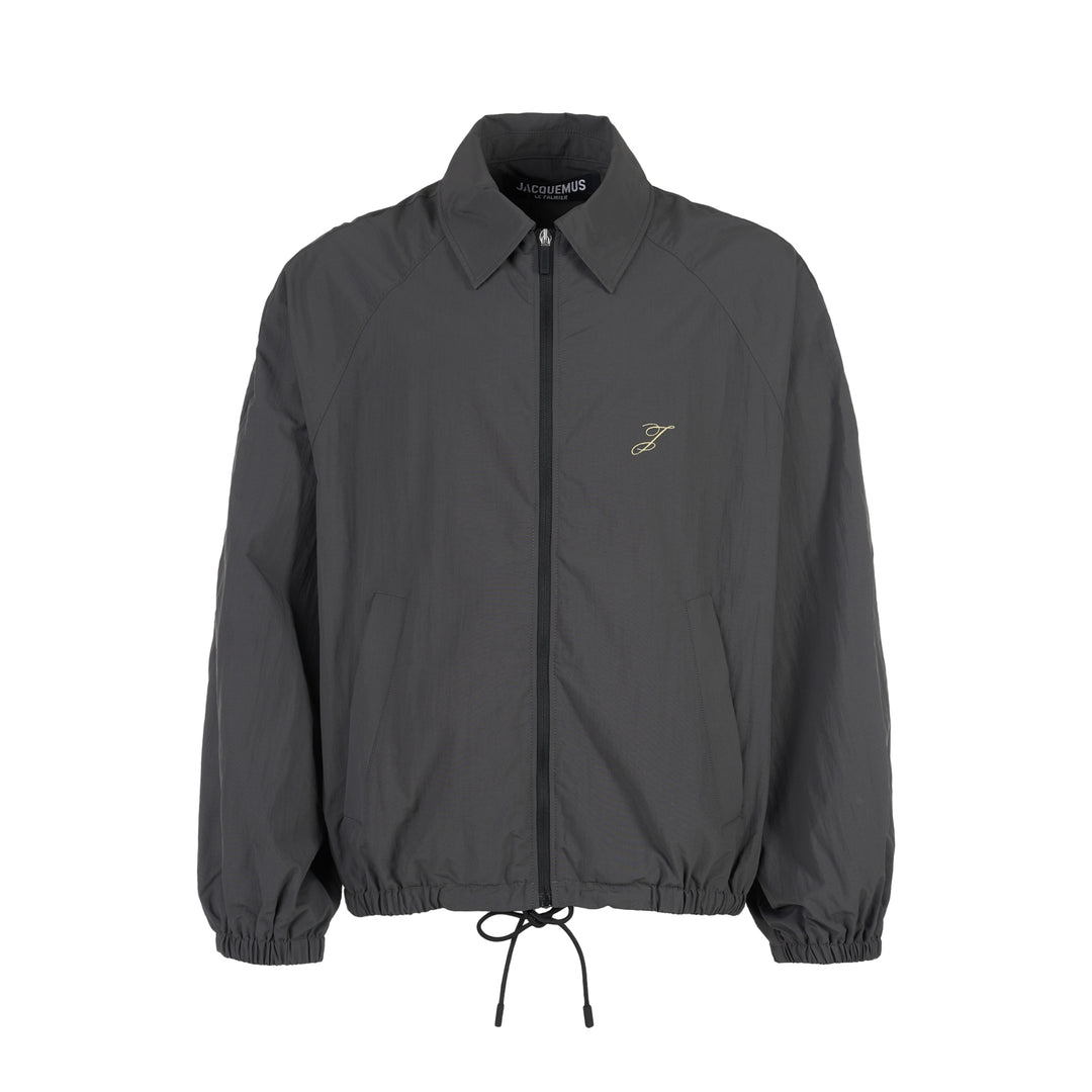 Jacquemus Le Coupe-Vent charcoal grey zip-up windbreaker with collar, elasticated cuffs and hem, and embroidered gold monogram on the chest