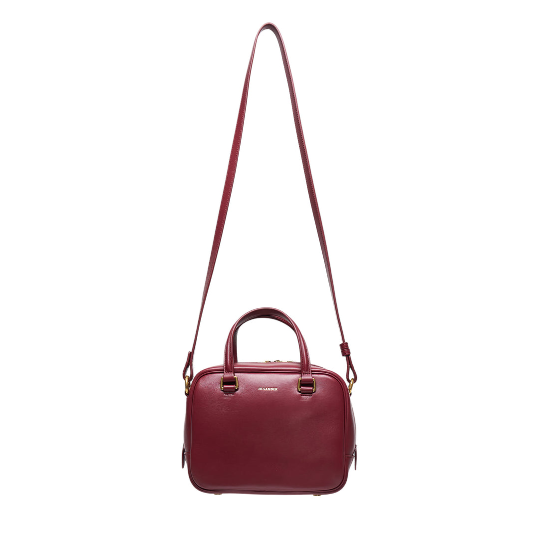 Jil Sander Vision Small burgundy nappa lambskin bowling bag with dual top handles, adjustable crossbody strap, and gold-tone hardware on a white background
