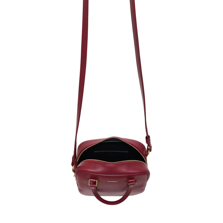 Jil Sander Vision Small burgundy nappa lambskin bowling bag with dual top handles, adjustable crossbody strap, and gold-tone hardware on a white background