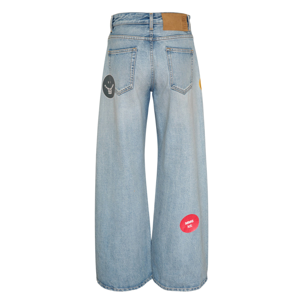 MM6 Maison Margiela wide-leg five-pocket jeans in light-wash blue denim, featuring eclectic sticker-style patches including a red heart, green MM6 diamond logo, circular branded roundels, and a hat-and-boot motif on the front panels