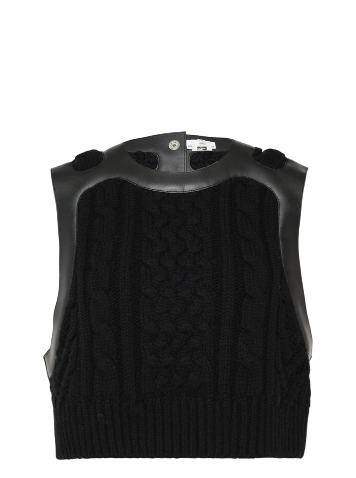 Noir Kei Ninomiya black cable-knit sleeveless vest with structured leather yoke, cutout detailing, and silver press-stud fastening at the rear neckline