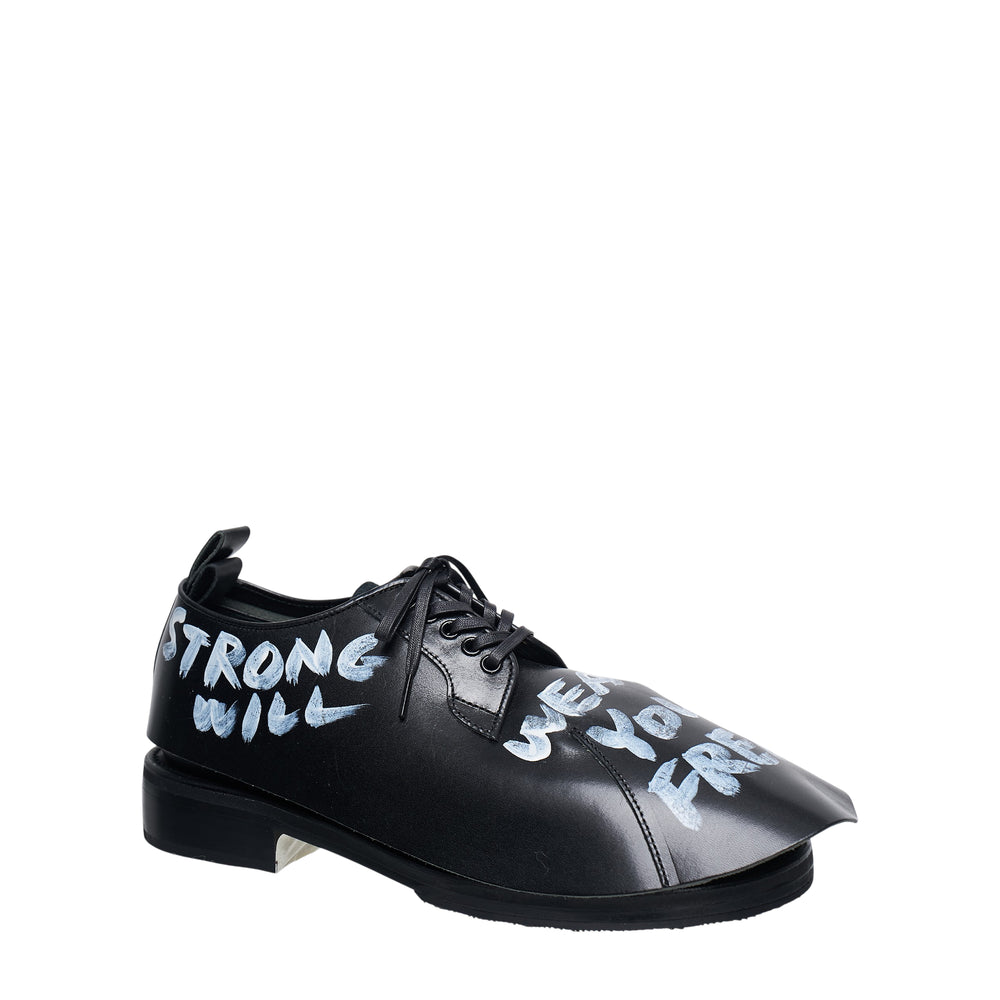 Comme des Garçons Homme Plus x Kids Love Gaite black leather oxford shoe with hand-painted white graffiti lettering reading 'Strong Will', featuring a sharply pointed toe and black ribbon laces, shown in profile view - view 2