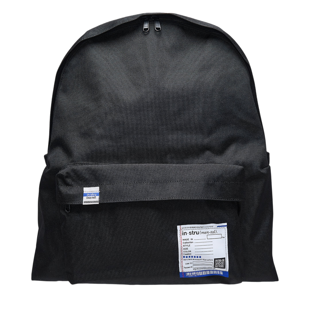 Mihara Yasuhiro in·stru(men·tal). large backpack in black canvas, front view showing zip closure, front pocket, woven brand label, and signature information-card patch with barcode and QR code detail