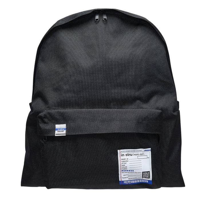 Mihara Yasuhiro in·stru(men·tal). large backpack in black canvas, front view showing zip closure, front pocket, woven brand label, and signature information-card patch with barcode and QR code detail