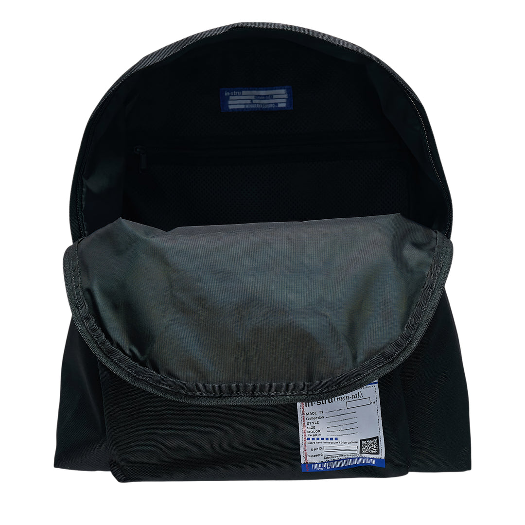 Mihara Yasuhiro in·stru(men·tal). large backpack in black canvas, front view showing zip closure, front pocket, woven brand label, and signature information-card patch with barcode and QR code detail