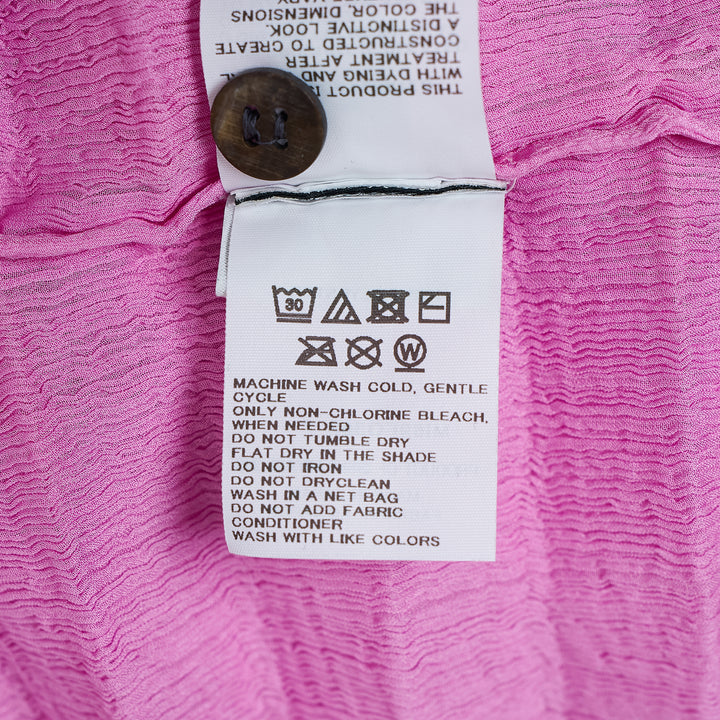me Issey Miyake Accordion See-Through Crepe Cardigan in pink, featuring horizontal accordion pleating, ruffled lettuce-edge trim, dark contrast button-through front, and a relaxed long-sleeve silhouette