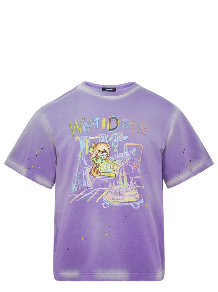 We11done Anniversary Teddy Graphic Tee in washed purple featuring a crayon-style teddy bear birthday illustration, multicolour logo lettering, and paint splatter details on an oversized cotton silhouette