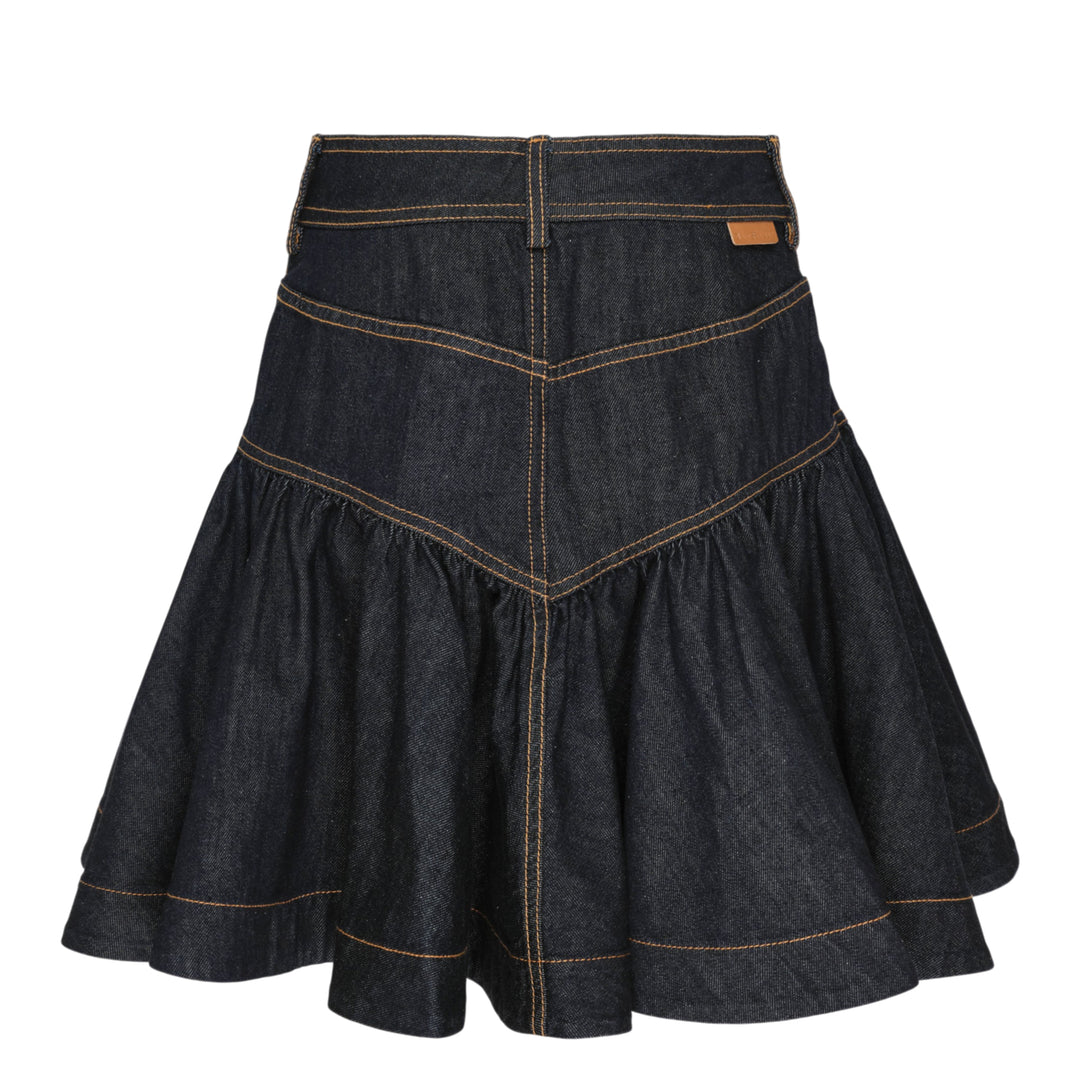 Zimmermann Denim Frill Mini Skirt in dark indigo with orange contrast topstitching, chevron seam detail, front patch pockets with branded gold snap buttons, and a gathered ruffle hem