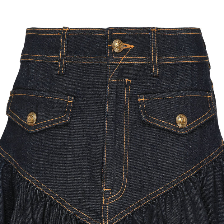 Zimmermann Denim Frill Mini Skirt in dark indigo with orange contrast topstitching, chevron seam detail, front patch pockets with branded gold snap buttons, and a gathered ruffle hem