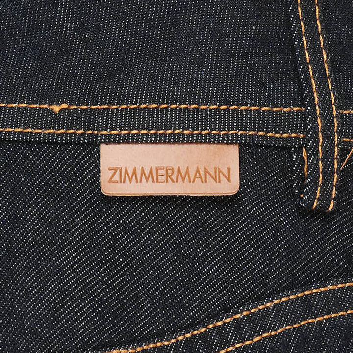 Zimmermann Denim Frill Mini Skirt in dark indigo with orange contrast topstitching, chevron seam detail, front patch pockets with branded gold snap buttons, and a gathered ruffle hem