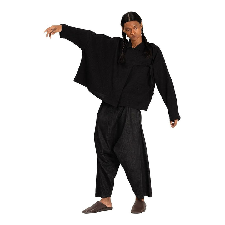 Model wearing Anaabu Asymmetrical Batwing Top in black textured fabric with an open collar, asymmetric front placket, patch chest pocket, and long batwing sleeves, styled with dark drop-crotch trousers and brown mules - packshot
