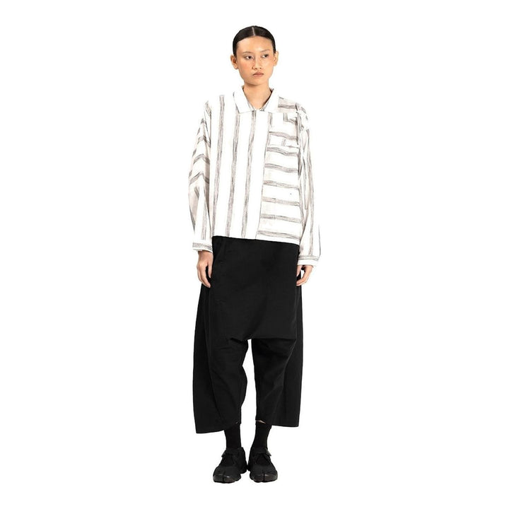 Model wearing Anaabu Asymmetrical Batwing Top in black textured fabric with an open collar, asymmetric front placket, patch chest pocket, and long batwing sleeves, styled with dark drop-crotch trousers and brown mules - packshot