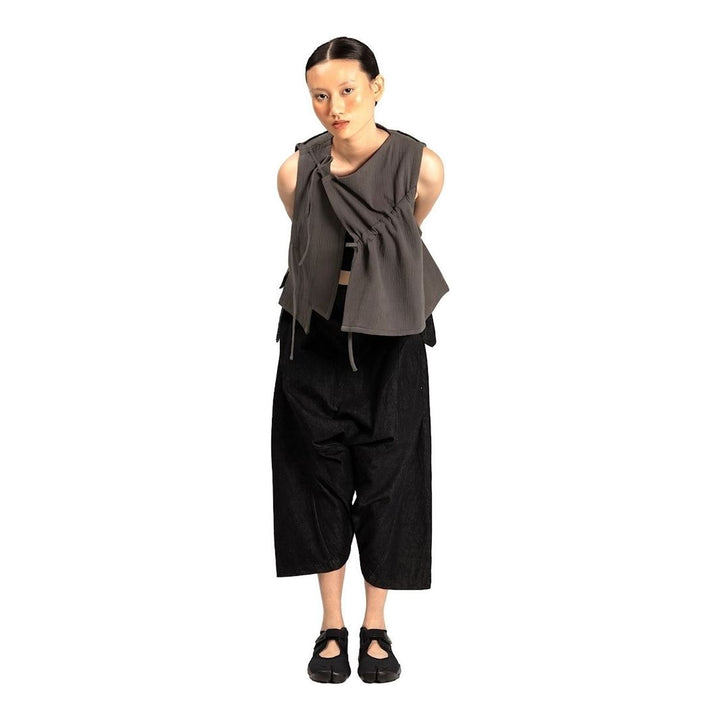 Model wearing the Anaabu Asymmetrical Vest in black, featuring an asymmetric neckline, diagonal panelling, and a draped fringe tie at the shoulder, styled with wide-leg black trousers and dark leather sandals. - packshot