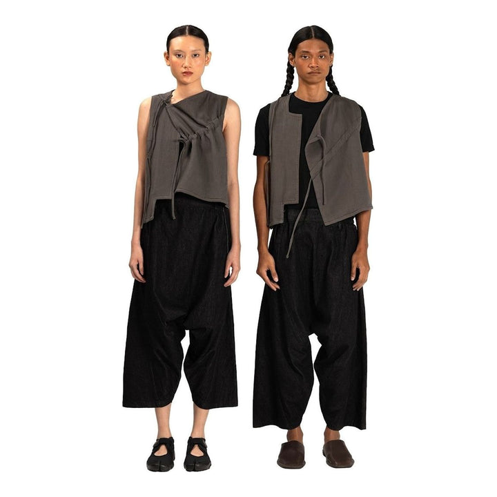 Model wearing the Anaabu Asymmetrical Vest in black, featuring an asymmetric neckline, diagonal panelling, and a draped fringe tie at the shoulder, styled with wide-leg black trousers and dark leather sandals. - packshot