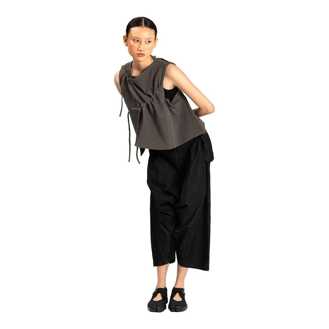 Model wearing the Anaabu Asymmetrical Vest in black, featuring an asymmetric neckline, diagonal panelling, and a draped fringe tie at the shoulder, styled with wide-leg black trousers and dark leather sandals. - packshot