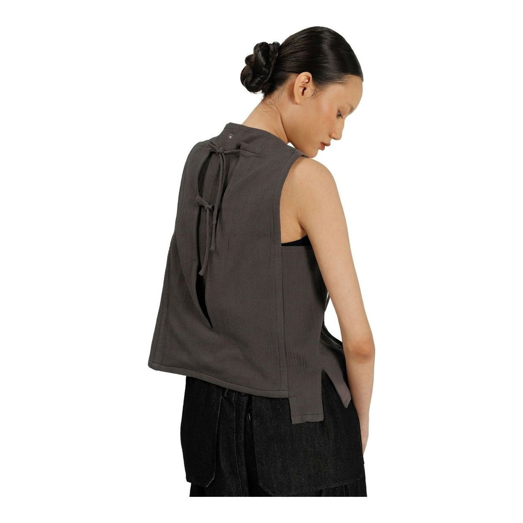 Model wearing the Anaabu Asymmetrical Vest in black, featuring an asymmetric neckline, diagonal panelling, and a draped fringe tie at the shoulder, styled with wide-leg black trousers and dark leather sandals. - packshot
