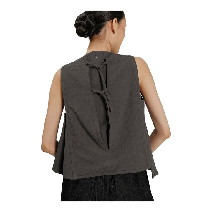 Model wearing the Anaabu Asymmetrical Vest in black, featuring an asymmetric neckline, diagonal panelling, and a draped fringe tie at the shoulder, styled with wide-leg black trousers and dark leather sandals. - packshot