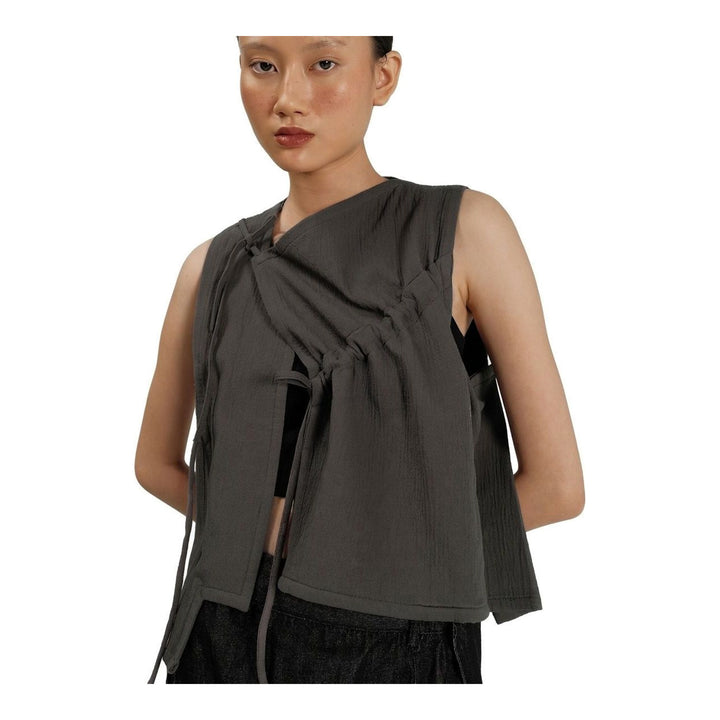 Model wearing the Anaabu Asymmetrical Vest in black, featuring an asymmetric neckline, diagonal panelling, and a draped fringe tie at the shoulder, styled with wide-leg black trousers and dark leather sandals. - packshot