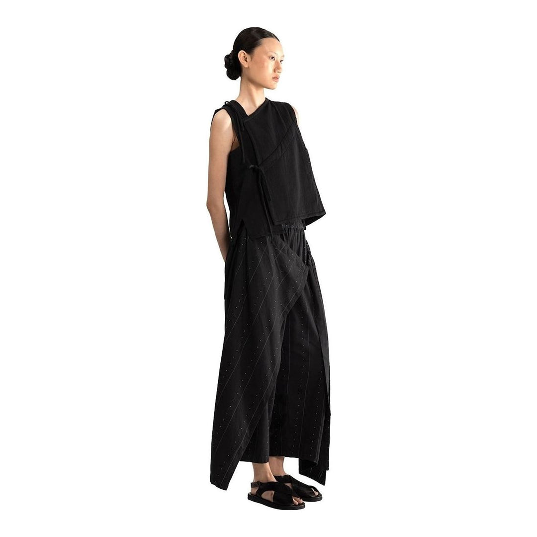 Model wearing the Anaabu Asymmetrical Vest in black, featuring an asymmetric neckline, diagonal panelling, and a draped fringe tie at the shoulder, styled with wide-leg black trousers and dark leather sandals. - packshot