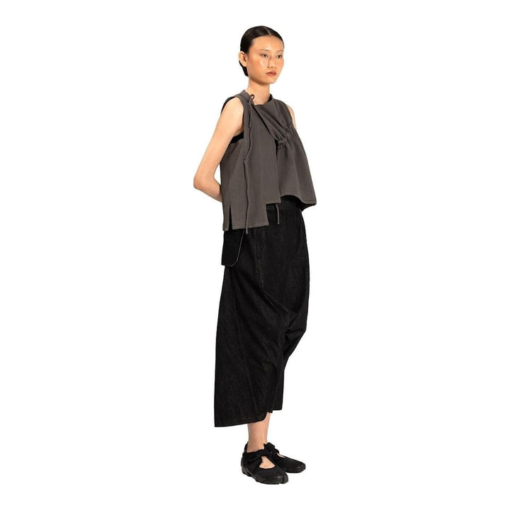 Model wearing the Anaabu Asymmetrical Vest in black, featuring an asymmetric neckline, diagonal panelling, and a draped fringe tie at the shoulder, styled with wide-leg black trousers and dark leather sandals. - packshot