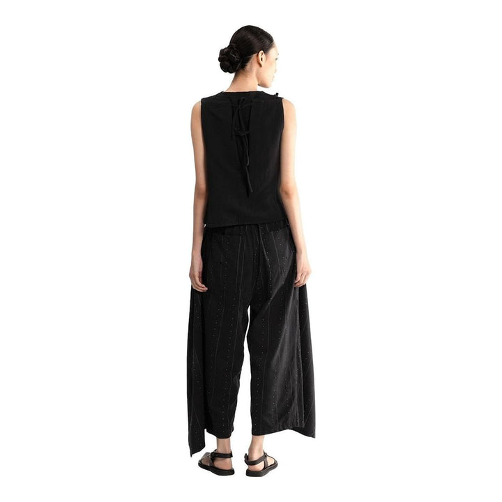 Model wearing the Anaabu Asymmetrical Vest in black, featuring an asymmetric neckline, diagonal panelling, and a draped fringe tie at the shoulder, styled with wide-leg black trousers and dark leather sandals. - packshot
