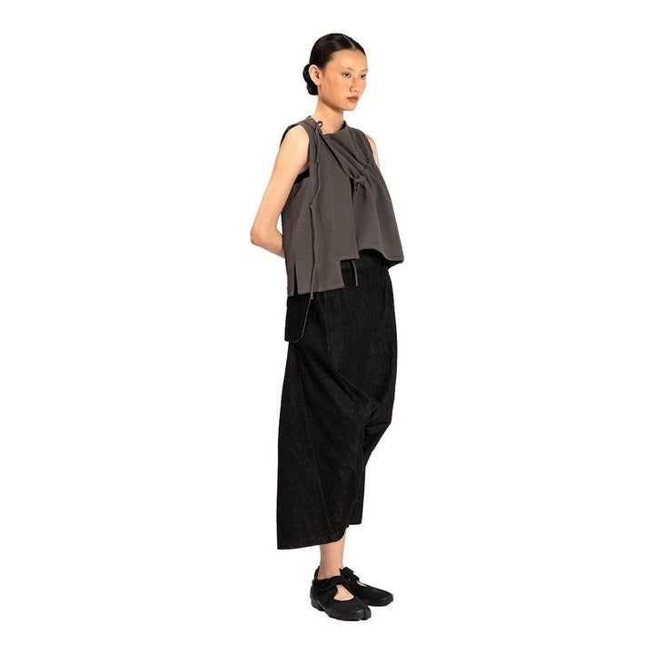 Model wearing the Anaabu Asymmetrical Vest in black, featuring an asymmetric neckline, diagonal panelling, and a draped fringe tie at the shoulder, styled with wide-leg black trousers and dark leather sandals. - packshot