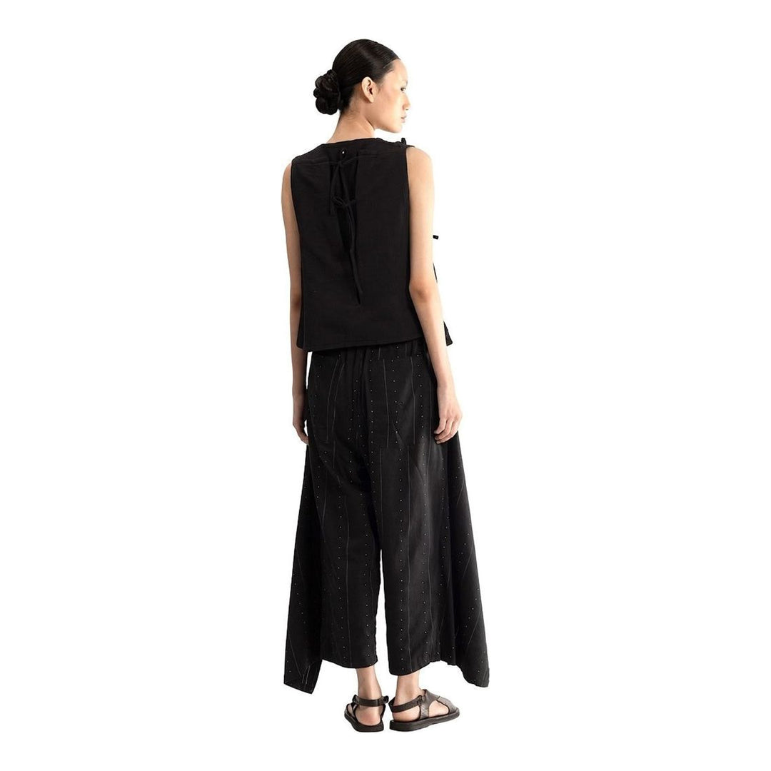 Model wearing the Anaabu Asymmetrical Vest in black, featuring an asymmetric neckline, diagonal panelling, and a draped fringe tie at the shoulder, styled with wide-leg black trousers and dark leather sandals. - packshot