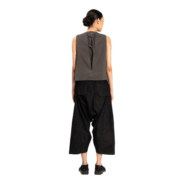 Model wearing the Anaabu Asymmetrical Vest in black, featuring an asymmetric neckline, diagonal panelling, and a draped fringe tie at the shoulder, styled with wide-leg black trousers and dark leather sandals. - packshot