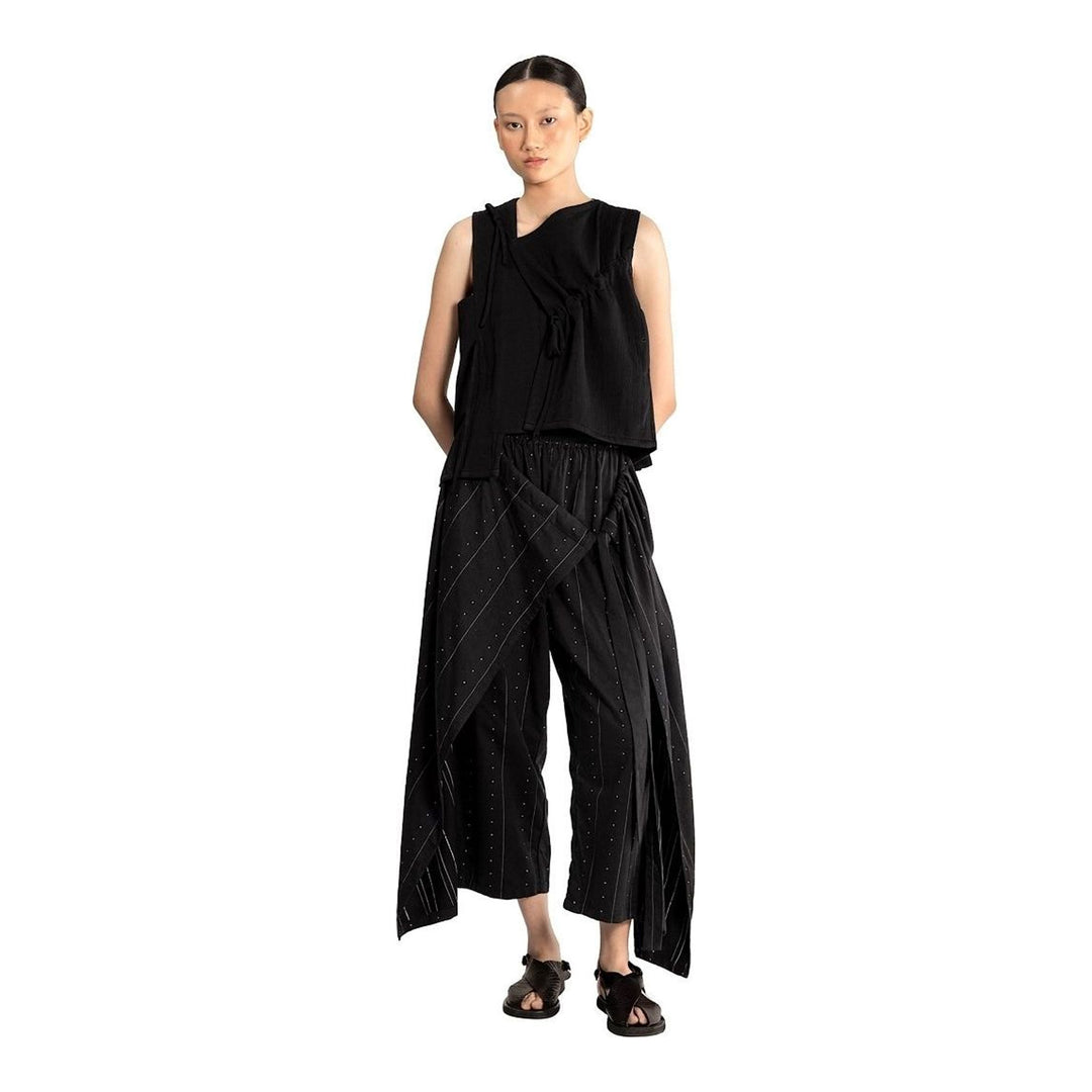 Model wearing the Anaabu Asymmetrical Vest in black, featuring an asymmetric neckline, diagonal panelling, and a draped fringe tie at the shoulder, styled with wide-leg black trousers and dark leather sandals. - packshot
