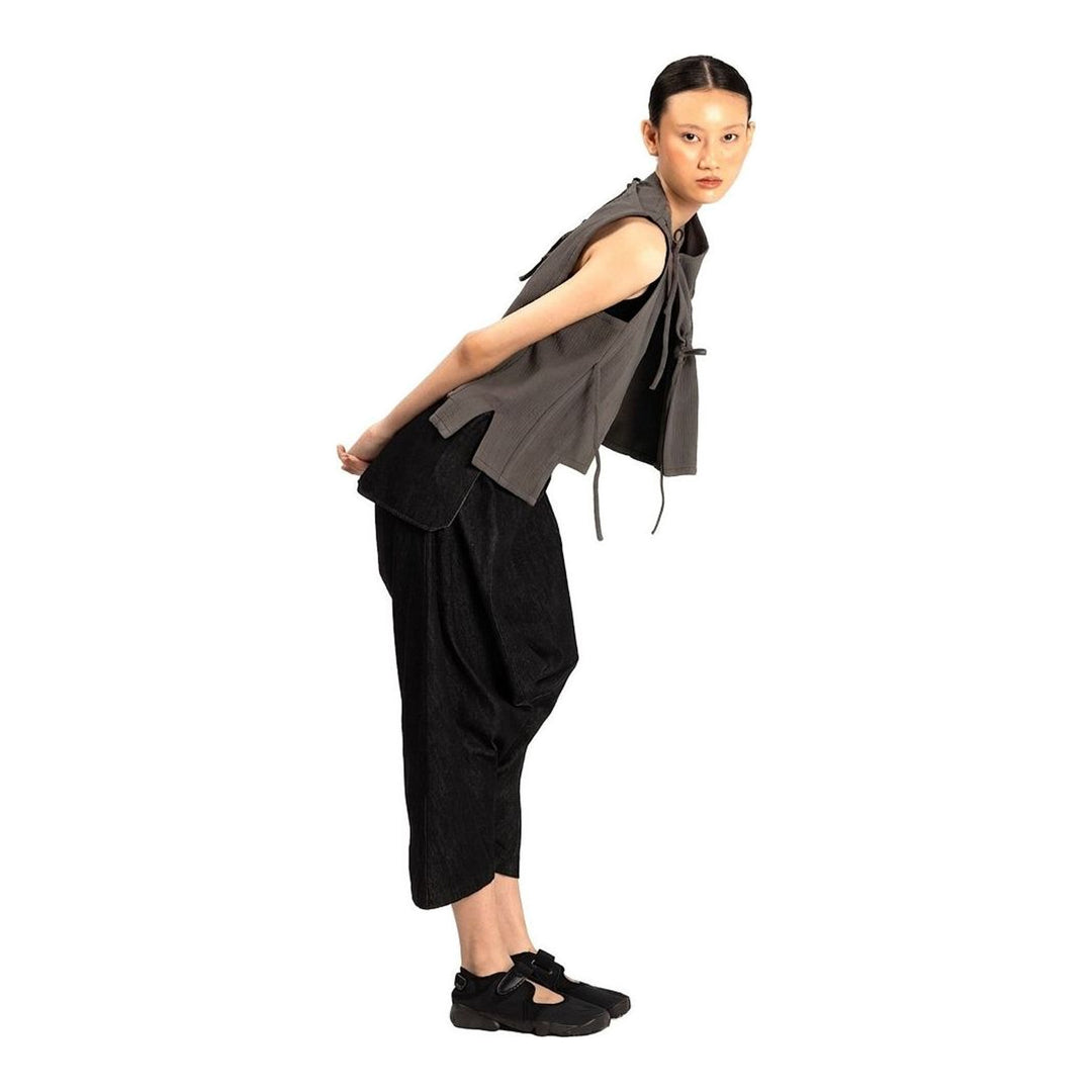 Model wearing the Anaabu Asymmetrical Vest in black, featuring an asymmetric neckline, diagonal panelling, and a draped fringe tie at the shoulder, styled with wide-leg black trousers and dark leather sandals. - packshot