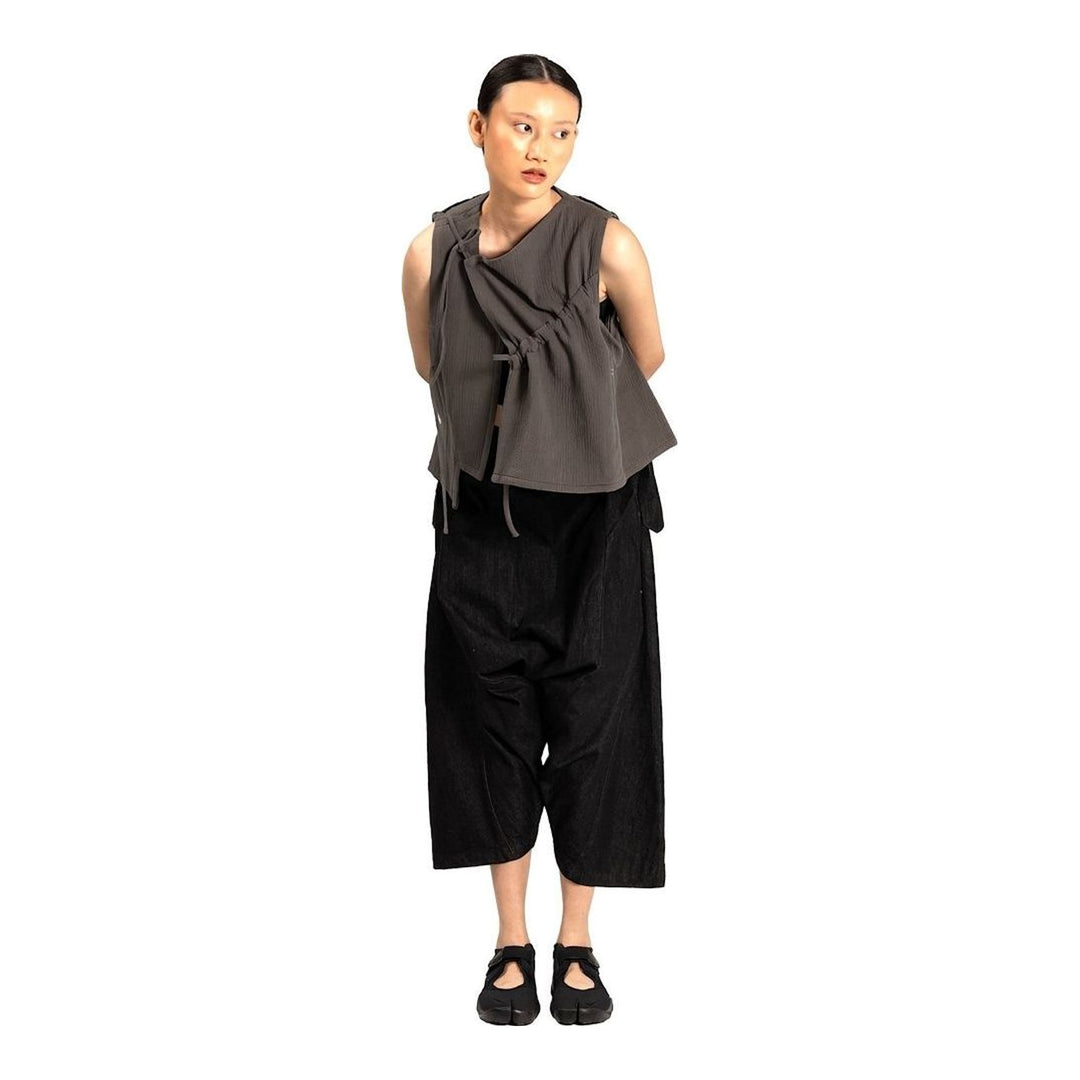 Model wearing the Anaabu Asymmetrical Vest in black, featuring an asymmetric neckline, diagonal panelling, and a draped fringe tie at the shoulder, styled with wide-leg black trousers and dark leather sandals. - packshot