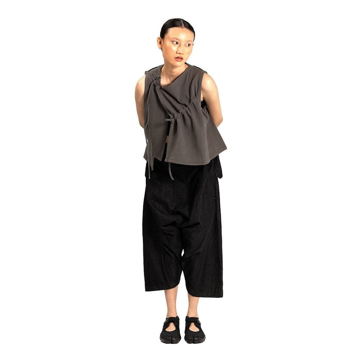 Model wearing the Anaabu Asymmetrical Vest in black, featuring an asymmetric neckline, diagonal panelling, and a draped fringe tie at the shoulder, styled with wide-leg black trousers and dark leather sandals. - packshot