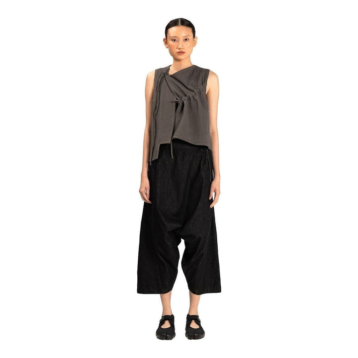 Model wearing the Anaabu Asymmetrical Vest in black, featuring an asymmetric neckline, diagonal panelling, and a draped fringe tie at the shoulder, styled with wide-leg black trousers and dark leather sandals. - packshot