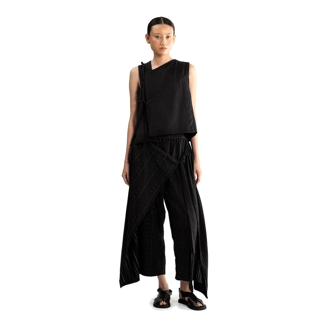 Model wearing the Anaabu Asymmetrical Vest in black, featuring an asymmetric neckline, diagonal panelling, and a draped fringe tie at the shoulder, styled with wide-leg black trousers and dark leather sandals. - packshot