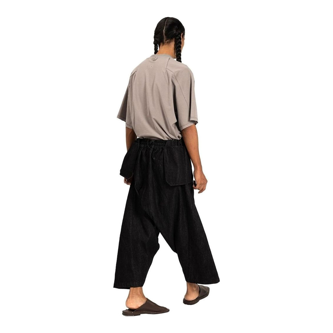 Model wearing Anaabu Dropped Culotte Pants in black — wide-leg drop-crotch trousers with an asymmetric draped skirt overlay panel, styled with a black crop top, frayed bucket hat, and tabi sneakers - packshot