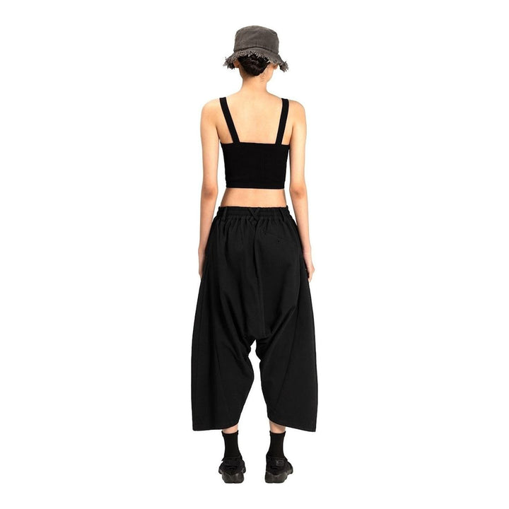 Model wearing Anaabu Dropped Culotte Pants in black — wide-leg drop-crotch trousers with an asymmetric draped skirt overlay panel, styled with a black crop top, frayed bucket hat, and tabi sneakers - packshot