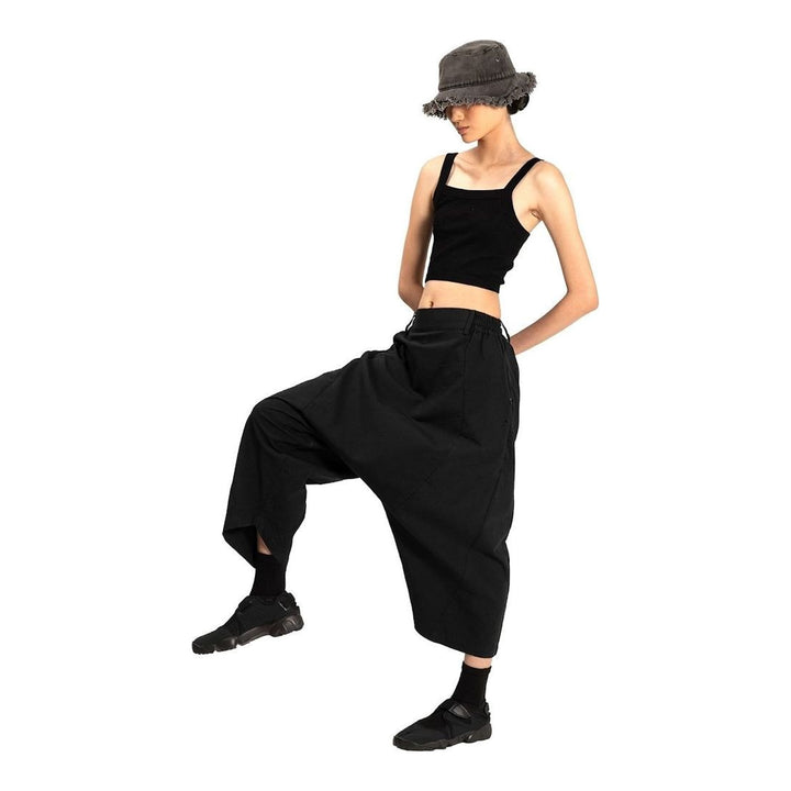 Model wearing Anaabu Dropped Culotte Pants in black — wide-leg drop-crotch trousers with an asymmetric draped skirt overlay panel, styled with a black crop top, frayed bucket hat, and tabi sneakers - packshot