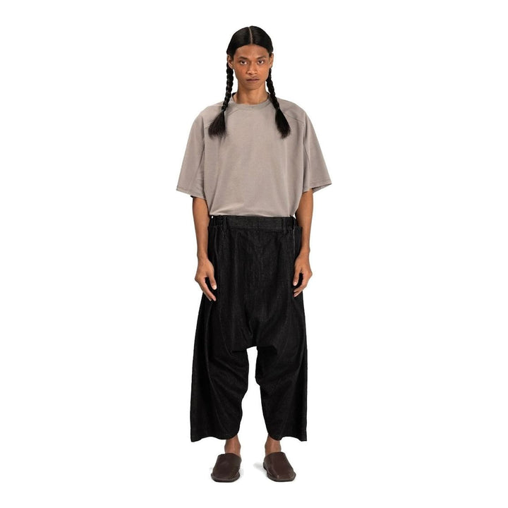 Model wearing Anaabu Dropped Culotte Pants in black — wide-leg drop-crotch trousers with an asymmetric draped skirt overlay panel, styled with a black crop top, frayed bucket hat, and tabi sneakers - packshot