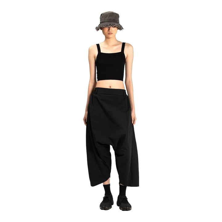 Model wearing Anaabu Dropped Culotte Pants in black — wide-leg drop-crotch trousers with an asymmetric draped skirt overlay panel, styled with a black crop top, frayed bucket hat, and tabi sneakers - packshot