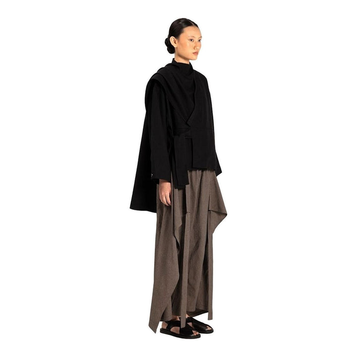 Model wearing the Anaabu Shawl Jacket in black — a draped wrap-style jacket with oversized shawl collar and self-tie belt, styled over a black turtleneck and olive wide-leg trousers - packshot