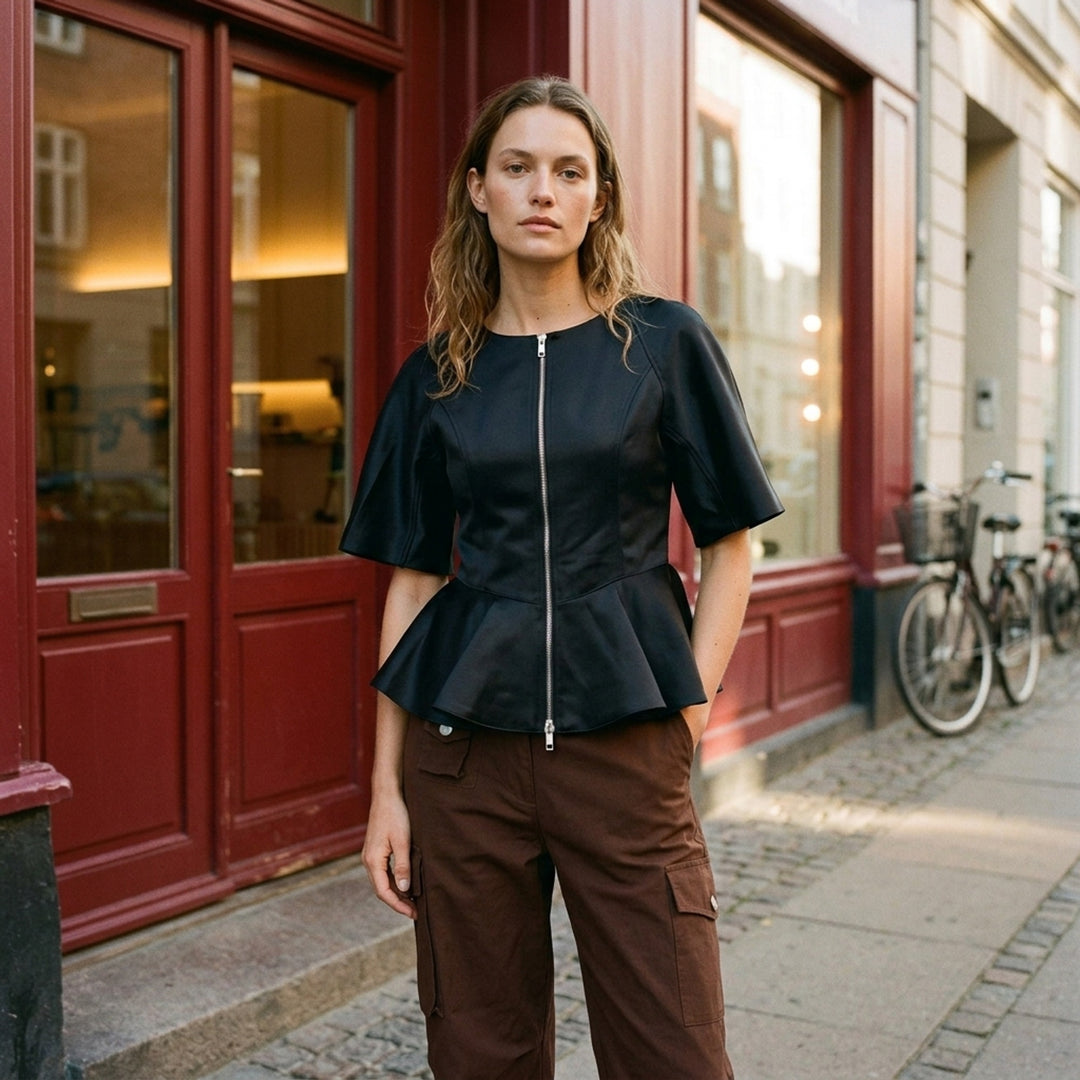 Ganni Duchesse Satin Peplum Zip Blouse in deep brown, displayed flat, featuring a full-length two-way zip, structured flared half-sleeves, and a dramatic fluted peplum hem - lifestyle