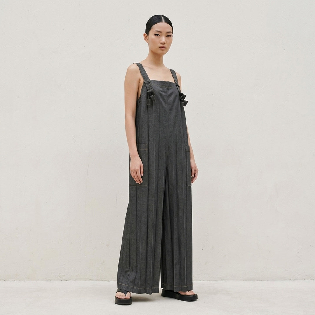 me Issey Miyake Denim Pleats Bottom jumpsuit in raw indigo denim, featuring deep vertical pleats, a square bib, knotted bow shoulder straps, patch hip pockets, and golden contrast topstitching - lifestyle