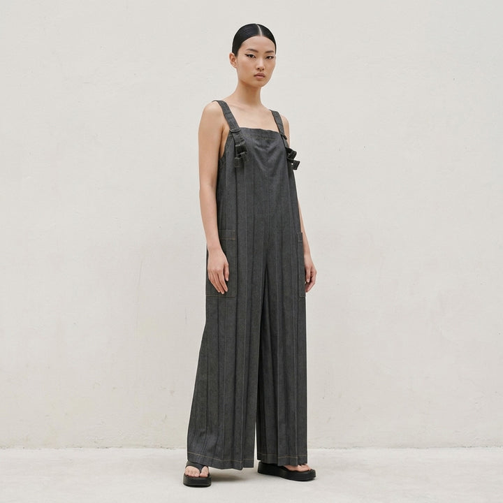 me Issey Miyake Denim Pleats Bottom jumpsuit in raw indigo denim, featuring deep vertical pleats, a square bib, knotted bow shoulder straps, patch hip pockets, and golden contrast topstitching - lifestyle