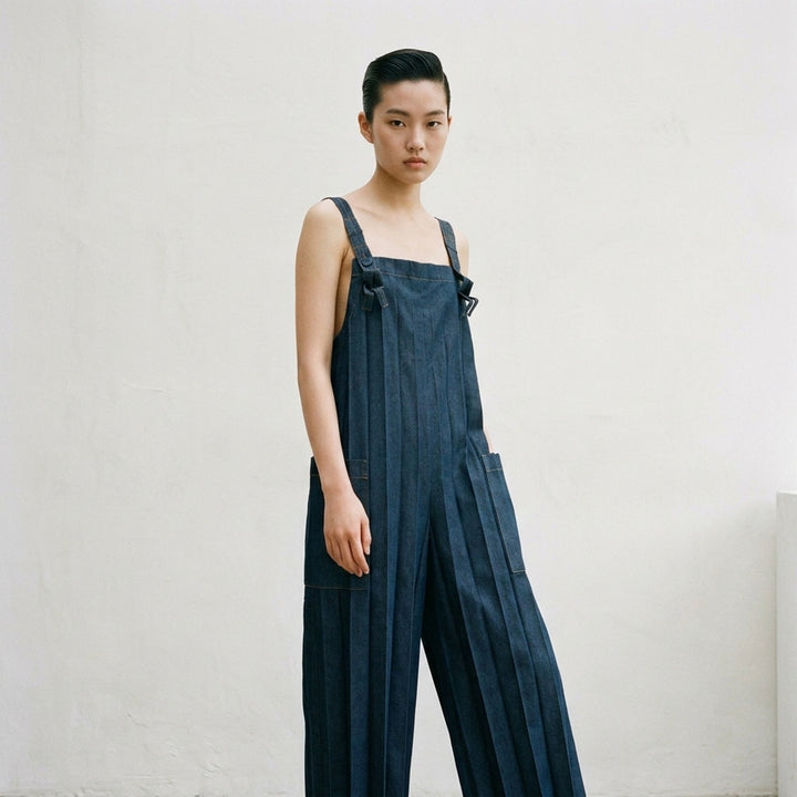 me Issey Miyake Denim Pleats Bottom jumpsuit in raw indigo denim, featuring deep vertical pleats, a square bib, knotted bow shoulder straps, patch hip pockets, and golden contrast topstitching - lifestyle